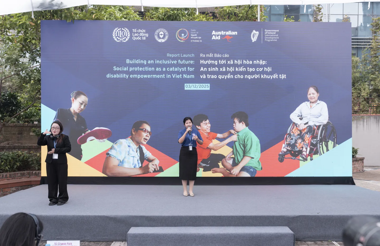 Vu Thi Hai Anh stands on stage speaking into a microphone at an ILO report launch event, with a sign language interpreter beside her and a large backdrop on inclusive social protection and disability empowerment behind them.