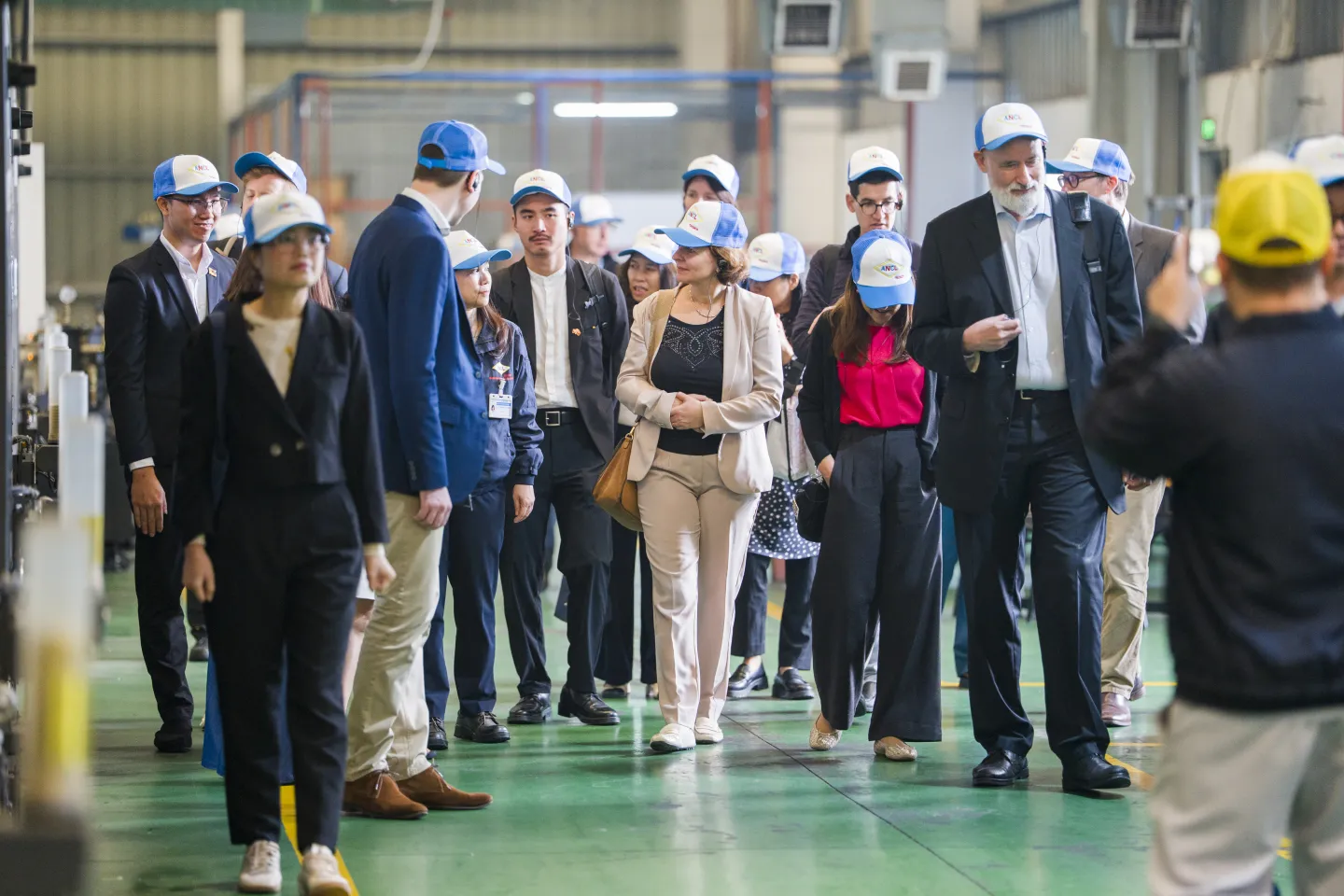 Members of the delegation walk through the factory floor during a site visit, accompanied by enterprise representatives, wearing safety caps while observing production areas.