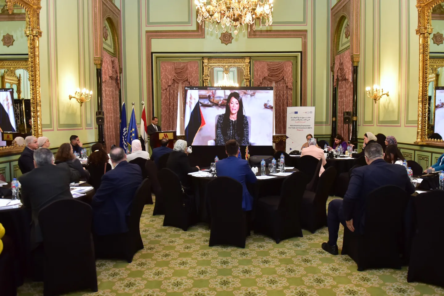 Meeting room with participants seated at round tables. On the screen plays a recorded message.