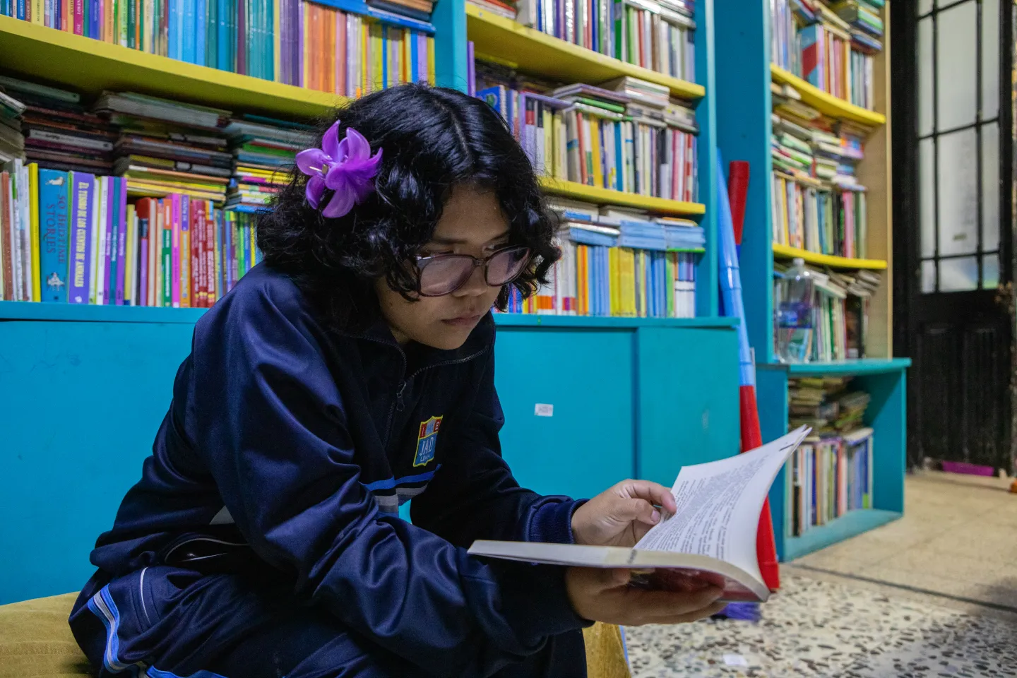 A teenage girl sits on a low bench inside a neighbourhood library, reading a book, with shelves filled with books behind her.