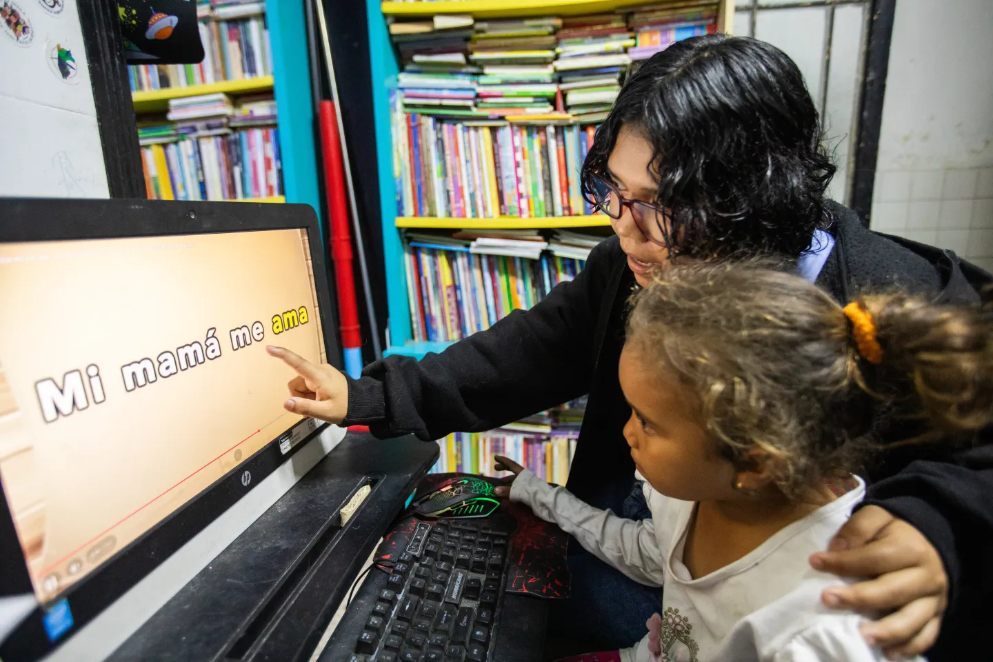 A teenage girl points at words on a computer screen while sitting next to a younger child inside a neighbourhood library, with bookshelves in the background.
