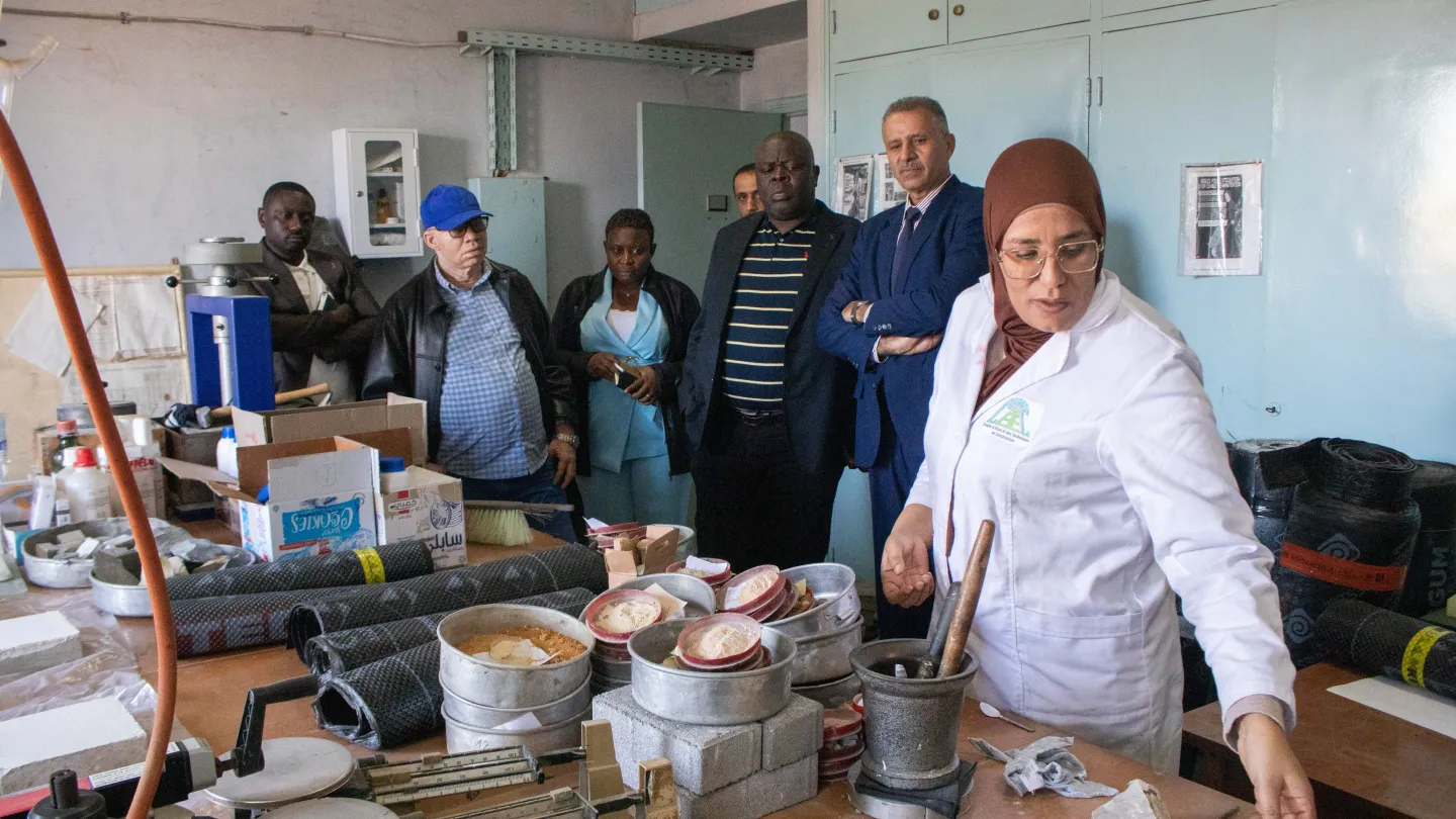 Technicienne en blouse de laboratoire présentant des équipements d’essai de matériaux de construction à un groupe de visiteurs dans un laboratoire.