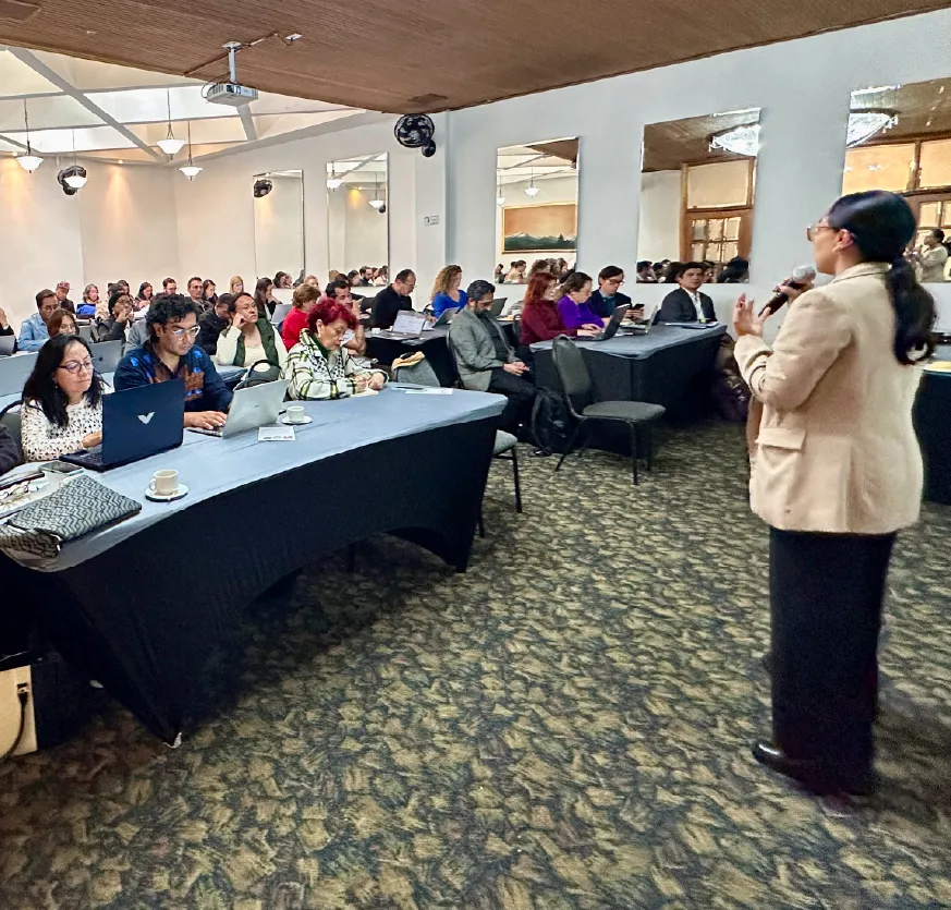 En un salón amplio tipo auditorio, varias filas de personas están sentadas frente a mesas largas, algunas con computadores portátiles y cuadernos. A la derecha, una mujer de pie habla con micrófono, dirigiéndose al público. El espacio es iluminado, con techo alto, espejos en las paredes y un ambiente de reunión formal o jornada de trabajo.