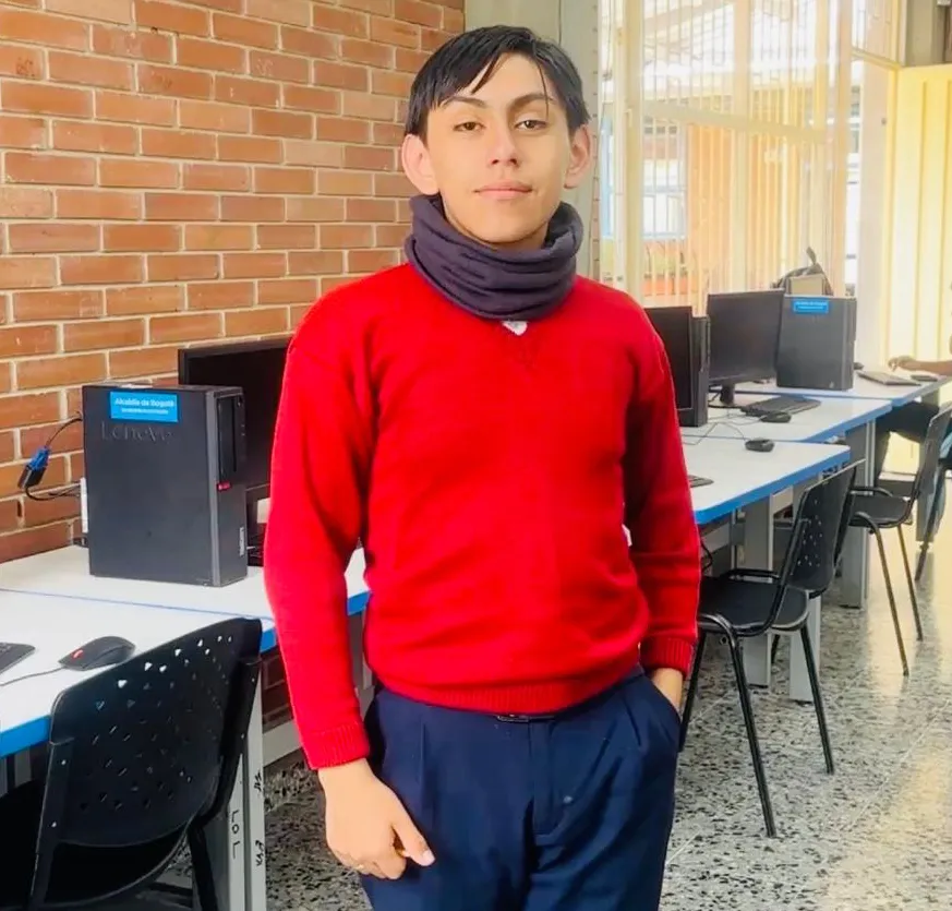 La imagen muestra a un adolescente de pie en una sala de informática de un colegio. El joven viste un suéter rojo de manga larga, pantalón oscuro y lleva una bufanda o cuello térmico oscuro alrededor del cuello. Tiene el cabello corto y oscuro, y mira de frente a la cámara con una expresión tranquila. Una de sus manos está en el bolsillo del pantalón