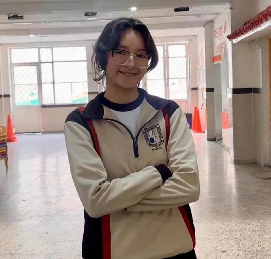  La imagen muestra a una adolescente de pie en el pasillo amplio de una institución educativa. La joven sonríe suavemente y tiene los brazos cruzados frente al cuerpo, lo que transmite seguridad y tranquilidad.