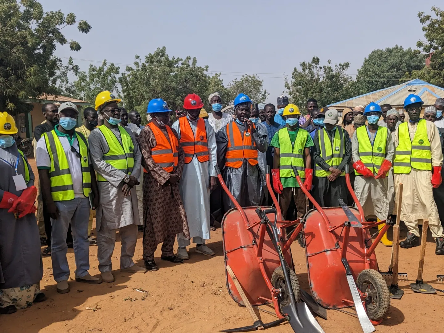 Group of officials and workers wearing hard hats and reflective vests stand outdoors during a flag-off event, with wheelbarrows and tools displayed in front of them.