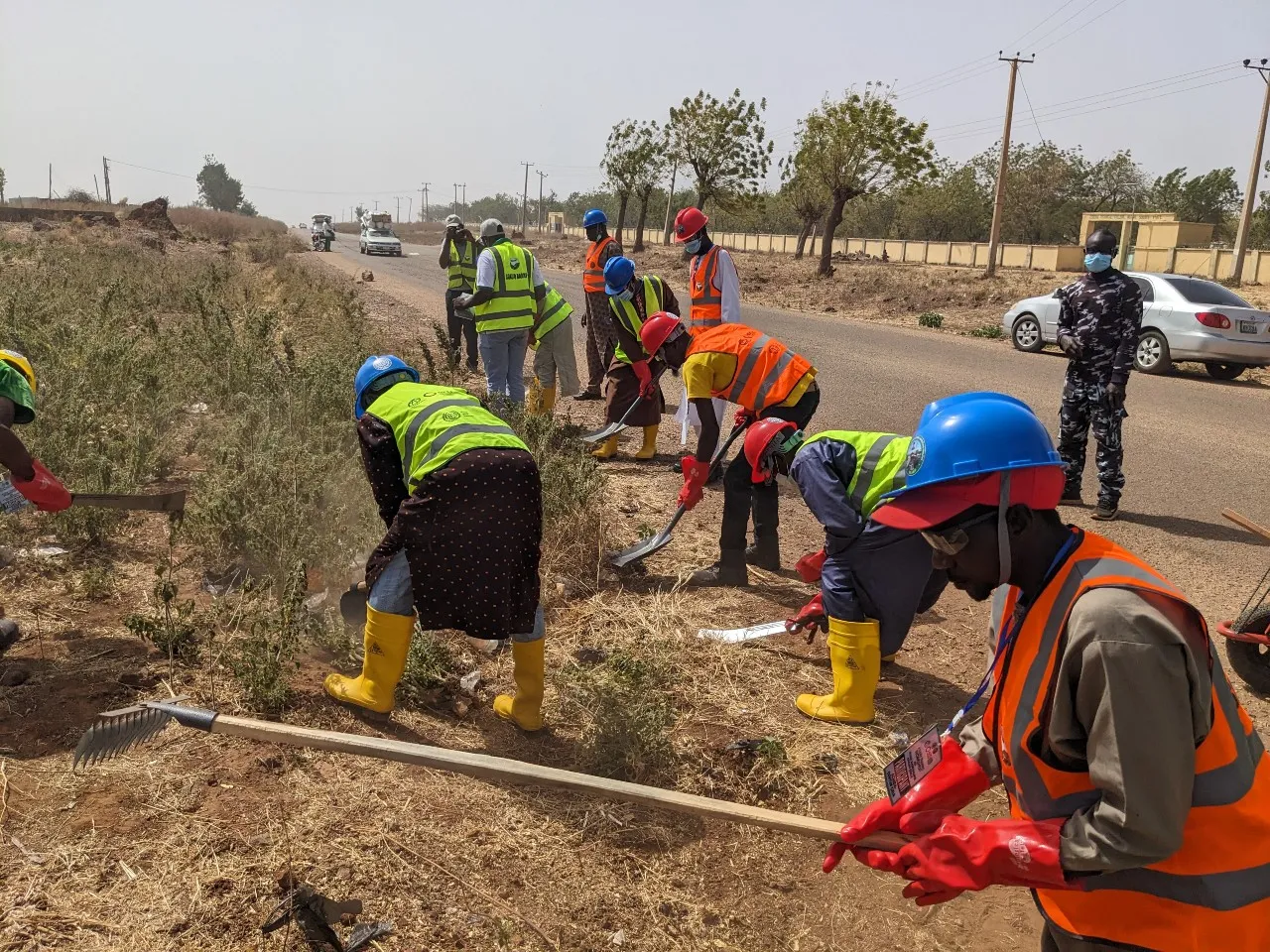 Workers wearing hard hats, reflective vests, gloves, and yellow boots clear roadside vegetation with hoes and rakes during the flag-off of routine maintenance works