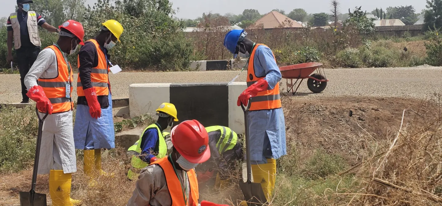 Five workers wearing hard hats, face masks, reflective safety vests, red gloves, and yellow rubber boots carry out hands-on fieldwork outdoors.