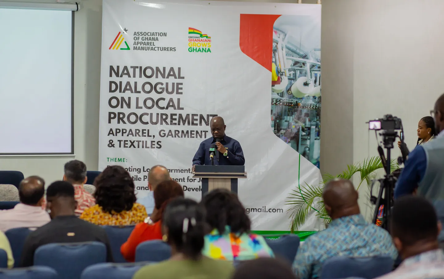 A man speaks at a podium in front of an audience during a conference on local garment production