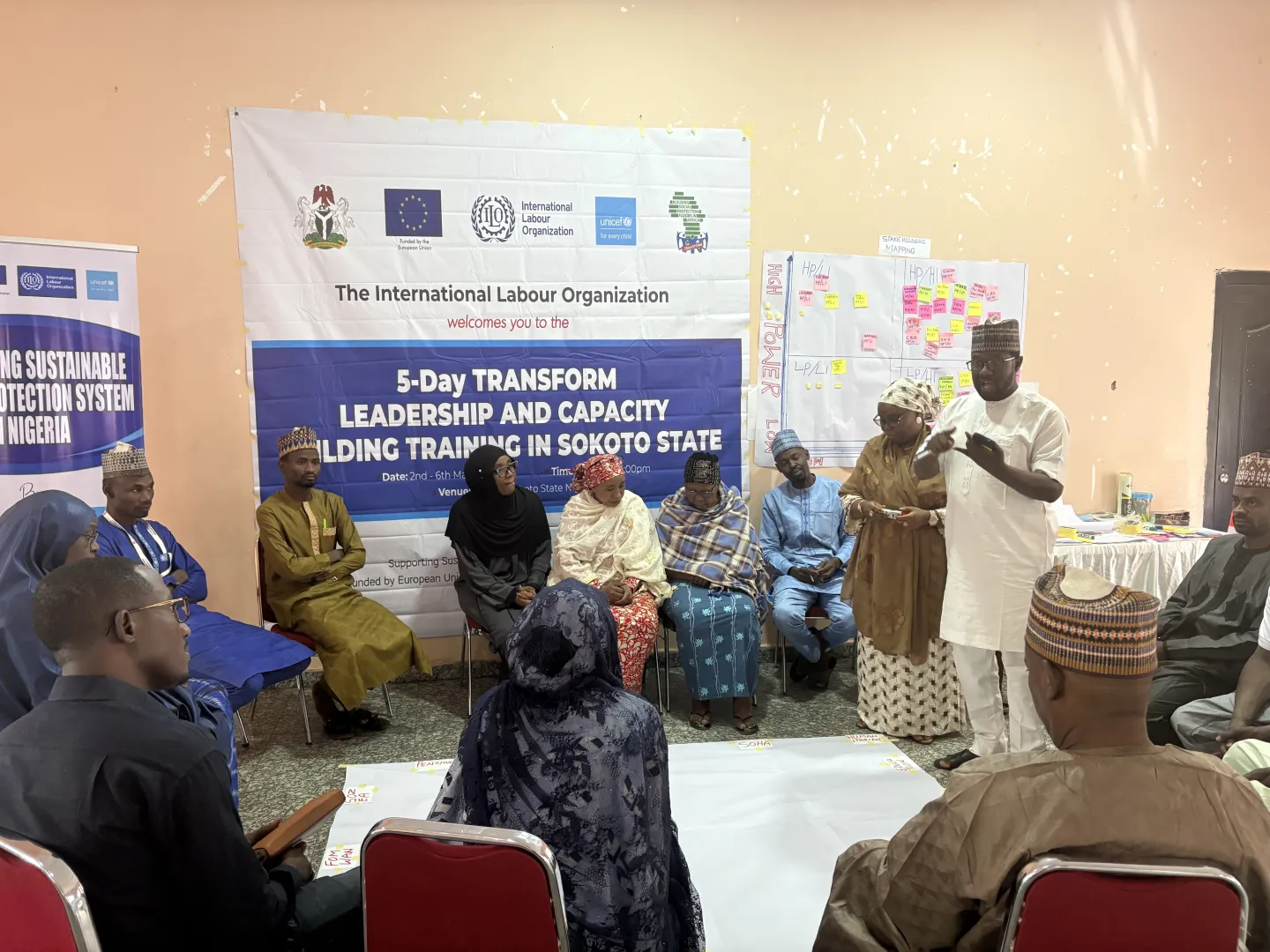 A group of men and women seated in a semi-circle while a facilitator stands and speaks, with a training banner displayed behind them.