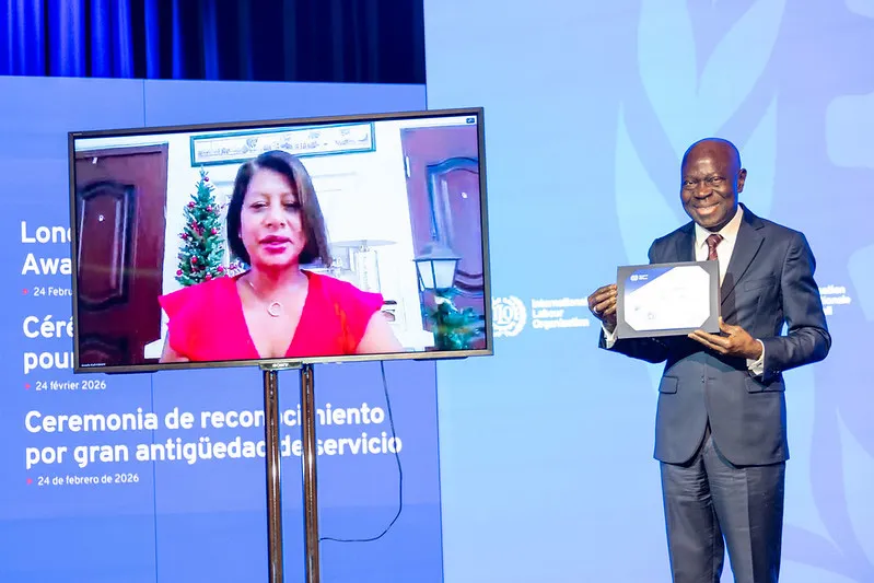 The ILO Director-General presents a Long Service Award certificate to Angela Colley, who participates remotely via video call and is displayed on screen beside him. 