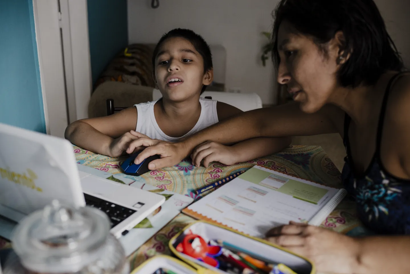 Una mujer ayuda a un niño con sus tareas frente a una computadora portátil. Ambos están sentados en una mesa con cuadernos abiertos, lápices de colores y otros útiles escolares. El niño utiliza el ratón mientras observa la pantalla, y la mujer lo acompaña de cerca, guiándolo en la actividad.