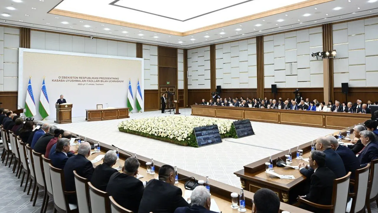 A conference room with officials seated around a large U-shaped table, listening to a speaker at a podium