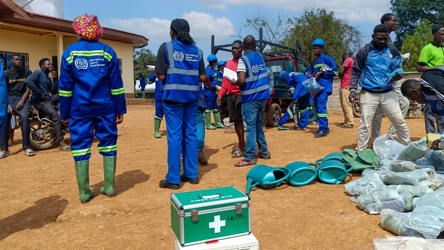 People in blue ILO work uniforms gather around protective equipment and supplies being distributed at an outdoor worksite.