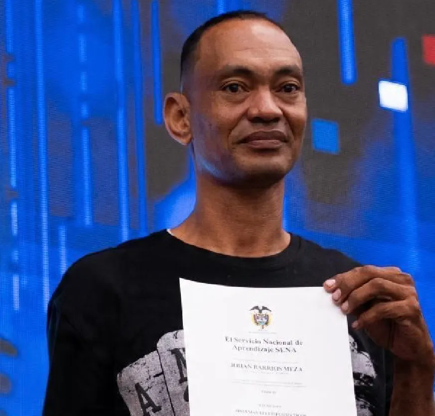 Un hombre adulto, contextura delgada y expresión serena está de pie frente a un fondo iluminado con tonos azules. Viste una camiseta negra y sostiene con ambas manos un diploma en posición vertical, a la altura del pecho. El documento corresponde al Servicio Nacional de Aprendizaje (SENA) y está a nombre de Johan Barrios Meza. El hombre mira a la cámara con una expresión que transmite orgullo y emoción. El entorno sugiere un escenario de evento o ceremonia, con iluminación profesional y un ambiente formal.