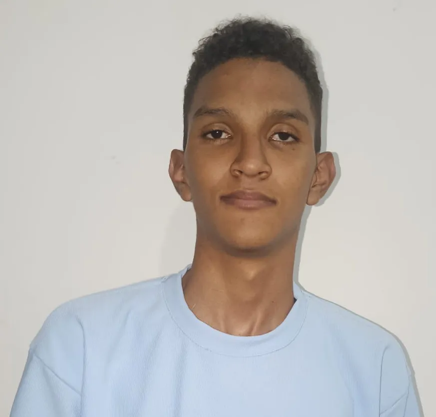 Un joven de pie posa frente a un fondo blanco. Está ubicado de frente a la cámara, con el cuerpo recto y los brazos relajados a los lados. Tiene el cabello corto, oscuro y ligeramente rizado, y una expresión serena, con la mirada directa hacia el lente. Viste una camiseta de color azul claro, de manga corta, sin estampados. Su contextura es delgada y su postura transmite tranquilidad y seguridad. 