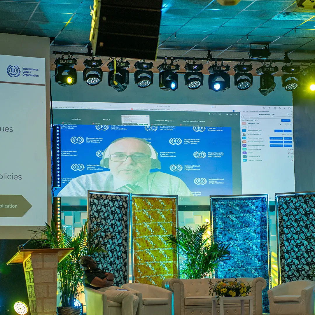 A photograph captures a large projection screen on a stage, showing a virtual presentation by Ariel Pino, ILO Specialist, Social Protection and Occupational Safety and Health during Annual General Assembly, National Tripartite Council (NTC) The Bahamas.