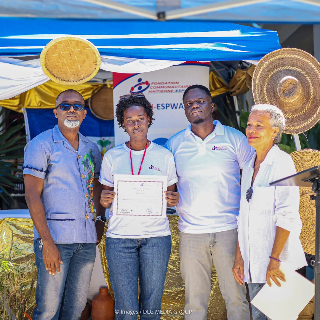 Four people stand together at an outdoor event under a decorated blue-and-white canopy. A young woman at the center holds a certificate while two men and an older woman stand beside her. Behind them are woven straw decorations, plants, and a banner