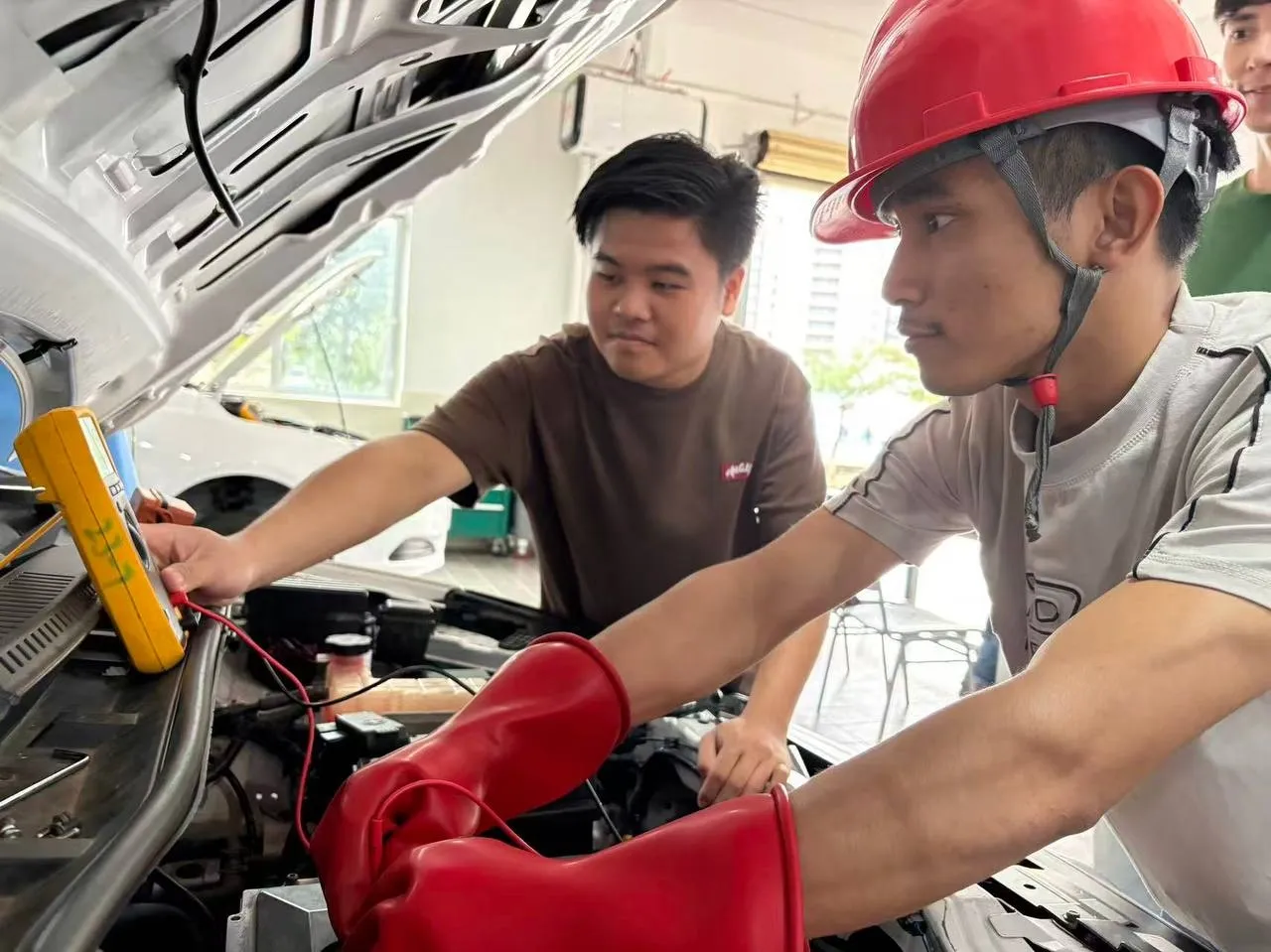 Two technicians inspect a vehicle, with one wearing a red safety helmet and both focusing on components under the vehicle’s hood.