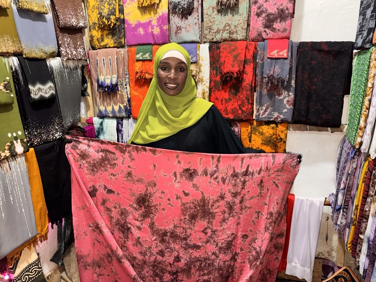 Semira Maye stands in her textile shop displaying one of her handmade fabrics, reflecting her journey toward rebuilding her livelihood through small business and entrepreneurship.