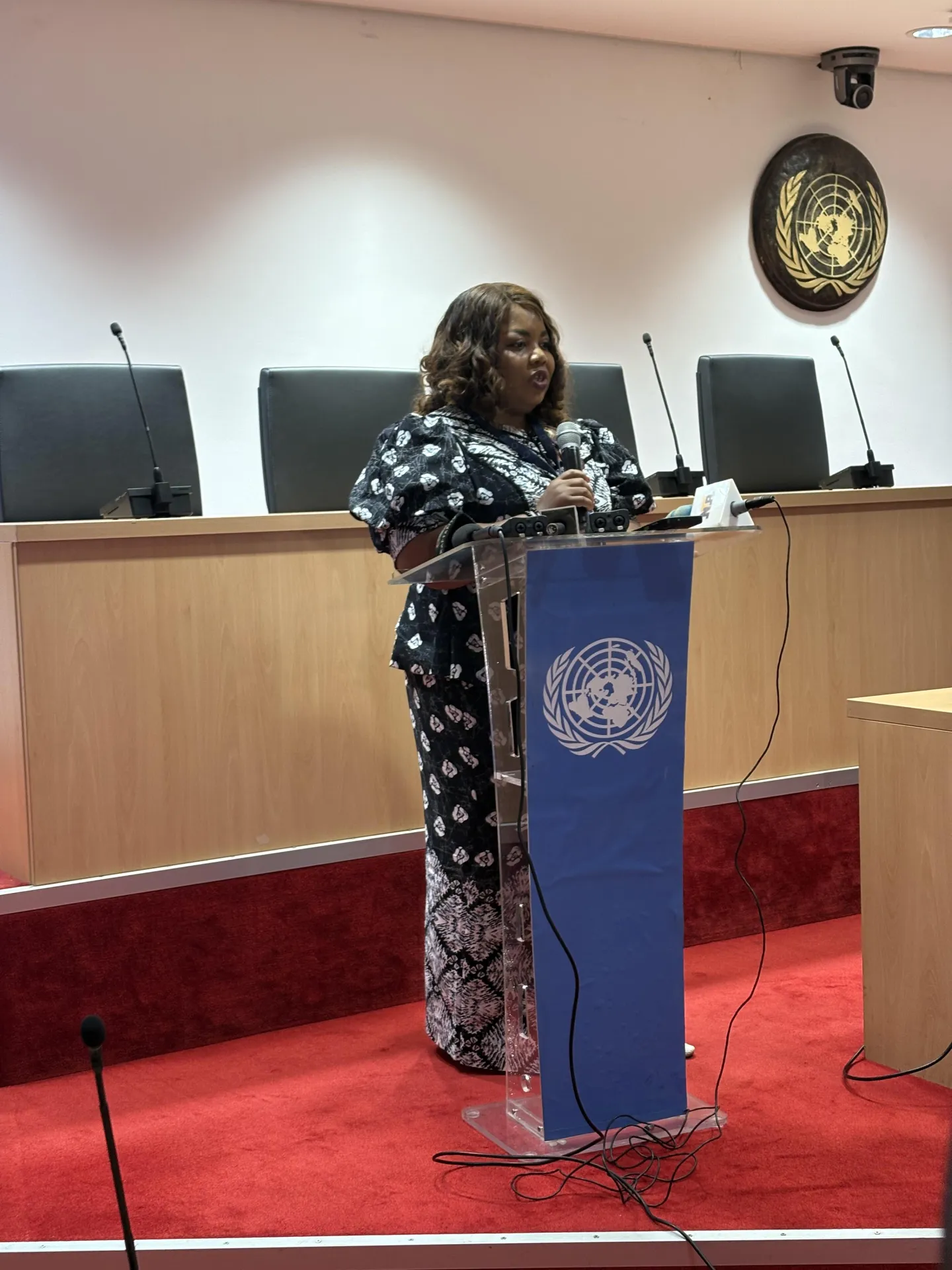 A woman speaking at a podium with a microphone inside a United Nations conference hall during an official event.