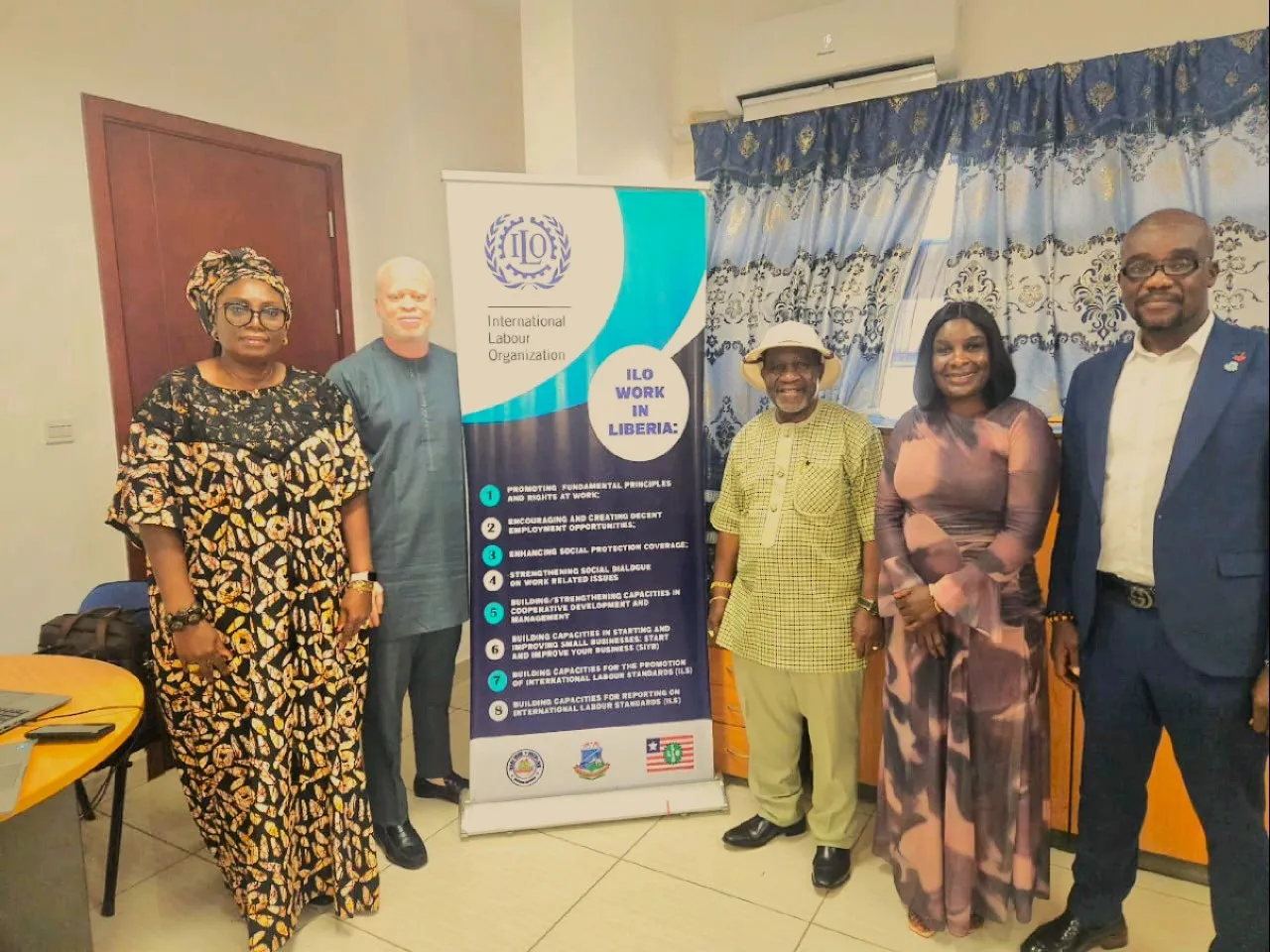 Five participants stand beside an International Labour Organization banner inside an office setting. The banner highlights ILO priorities in Liberia, including decent employment, labour standards, and social dialogue.