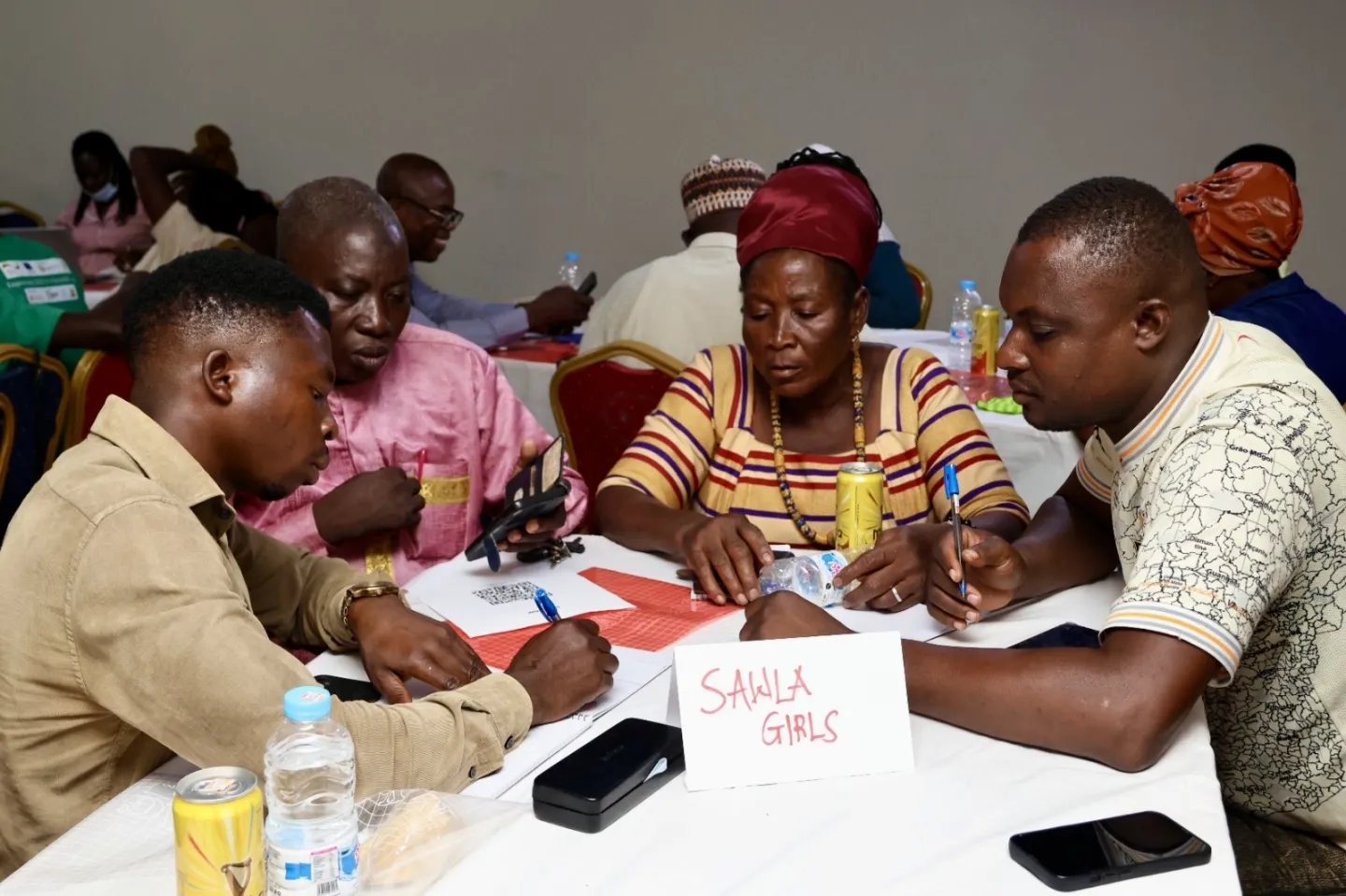 Four workshop participants seated around a table writing notes and discussing ideas during a training exercise. A sign labeled “Sawla Girls” is placed on the table alongside notebooks, phones, and water bottles.