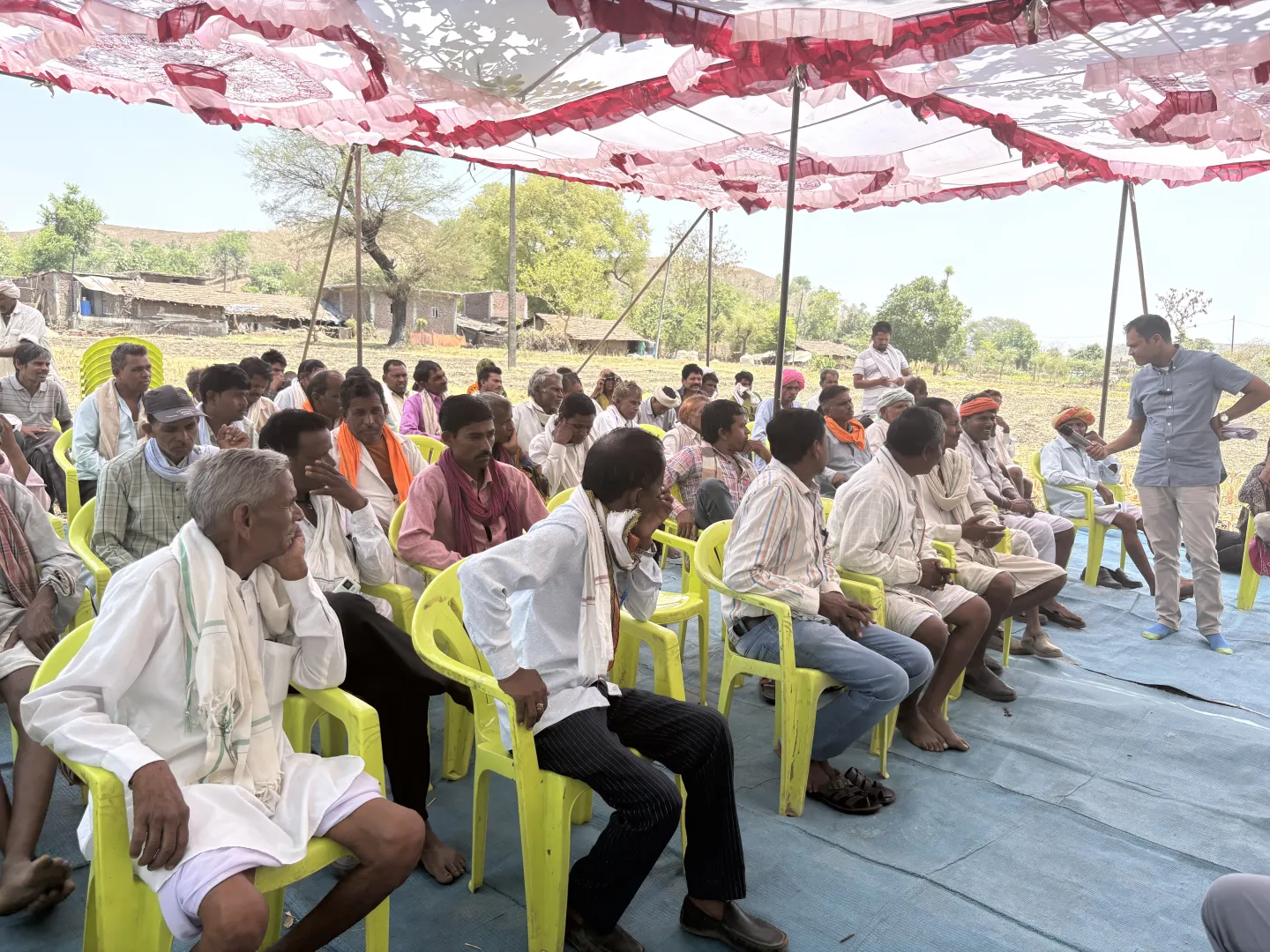 A large group of people seated in rows of chairs under a plastic sheet overhead.