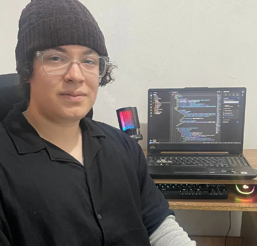 Un joven está sentado frente a un escritorio en un espacio interior de paredes claras. Mira directamente a la cámara con una expresión tranquila. Lleva un gorro tejido oscuro, gafas transparentes y una camiseta negra sobre una prenda de manga larga gris.