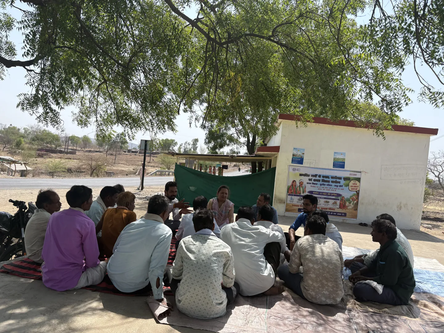 A large group of people sitting under the shade of a tree, listening to a speaker.