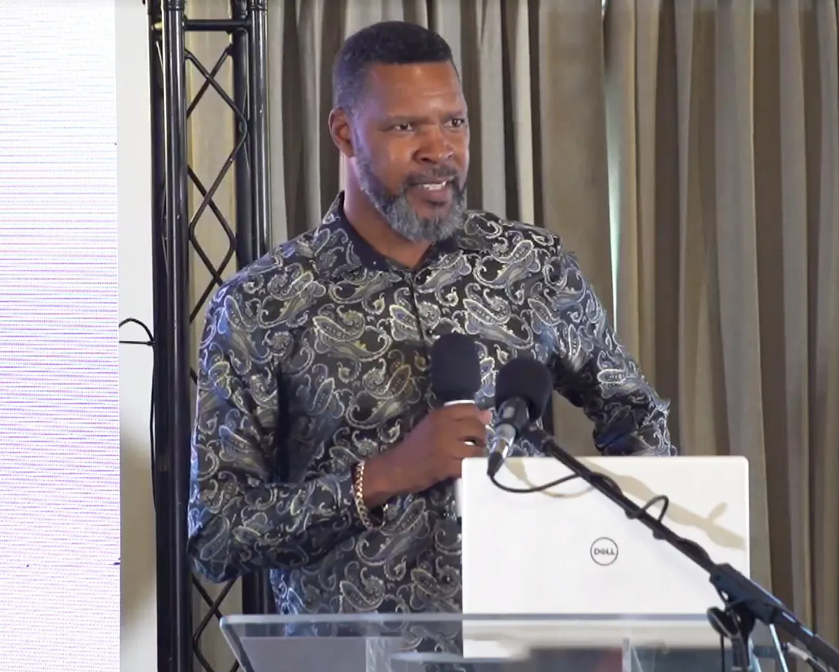 A man with a salt-and-pepper beard and a dark, paisley-patterned shirt speaking at a podium with microphones and a white laptop.