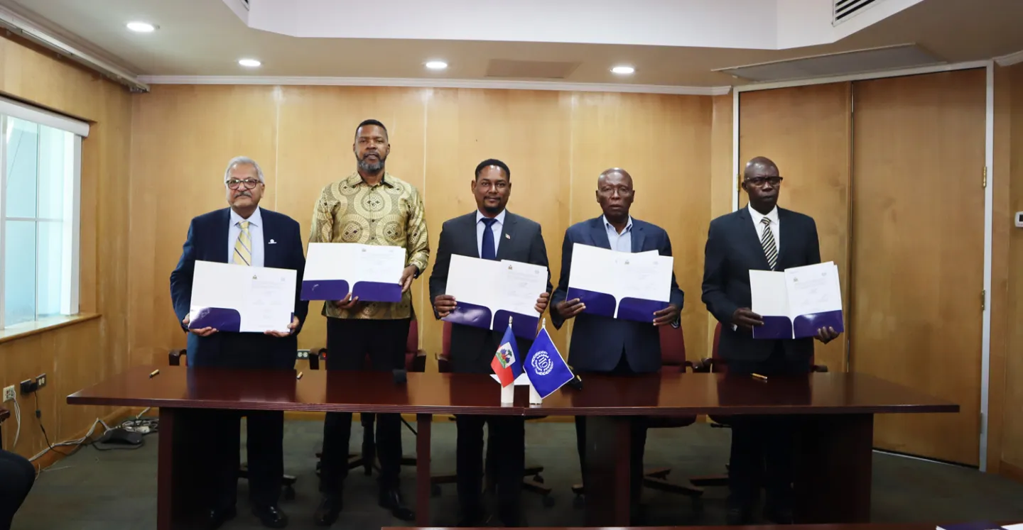 Five men stand side-by-side behind a long wooden conference table, each holding an open blue folder to display a signed document. Small desk flags for Haiti and the International Labour Organization (ILO) are positioned in the center of the table.