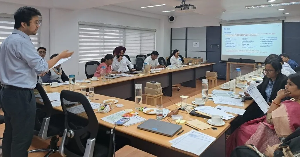 A man standing at the front of a training room speaks to a group of seated participants around a long conference table.