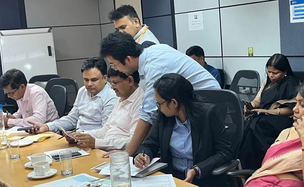 A group of people seated closely together at tables participate in a workshop activity. Some are writing or reading documents while others look on attentively, creating a focused and collaborative learning environment.