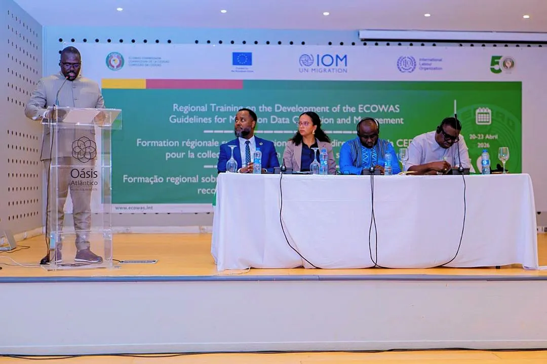 A speaker stands at a podium addressing participants seated at a panel during a regional migration data workshop in a conference hall with ECOWAS, EU, IOM, and ILO branding displayed behind them.