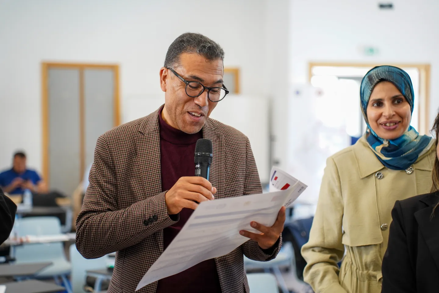 Un participant prend la parole avec un microphone lors d’un atelier, devant un groupe de participants assis autour de tables.