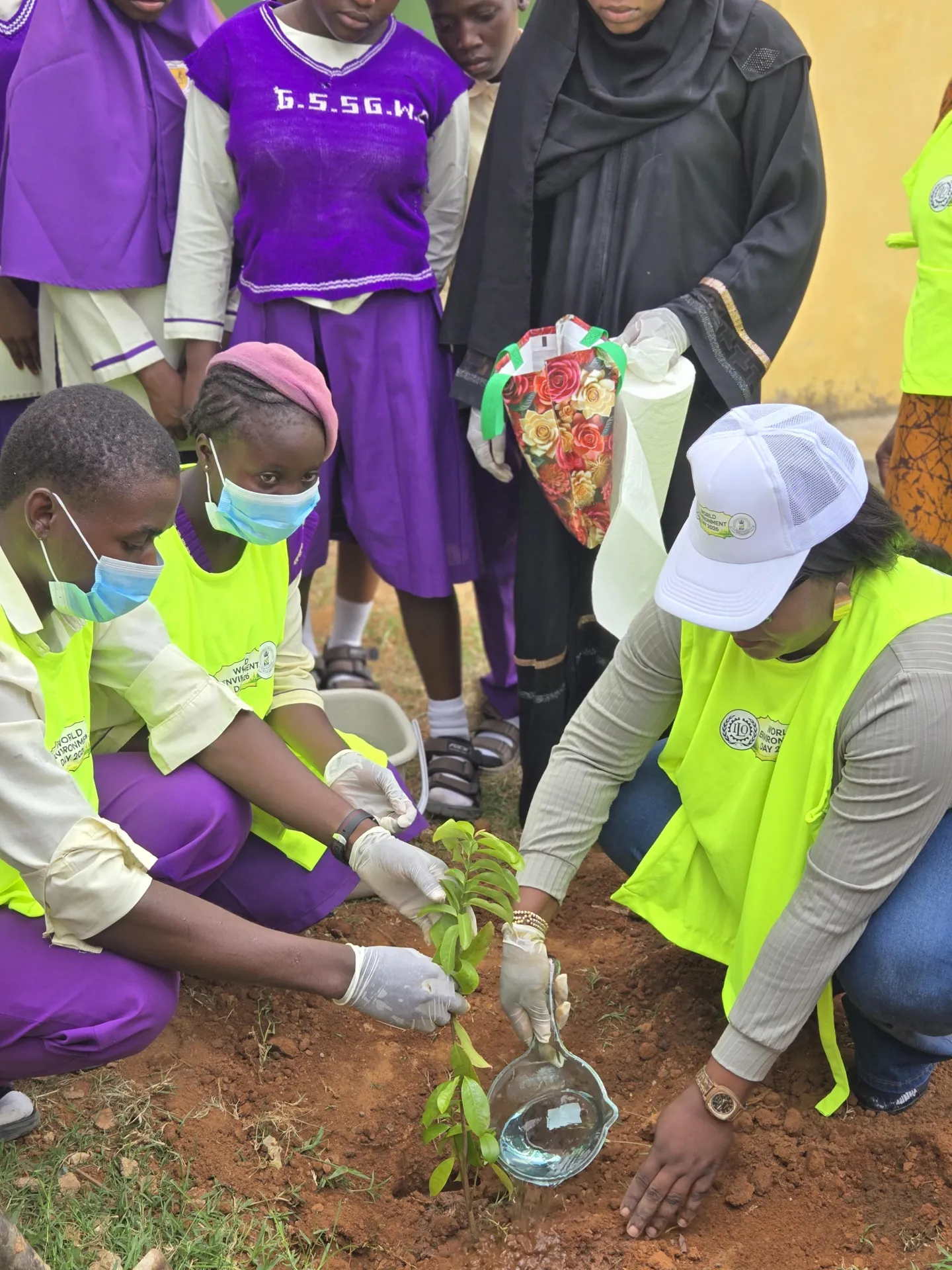 Students wearing school uniforms and high-visibility vests carefully plant and water a young tree while an official supervises, demonstrating practical climate action and environmental education.