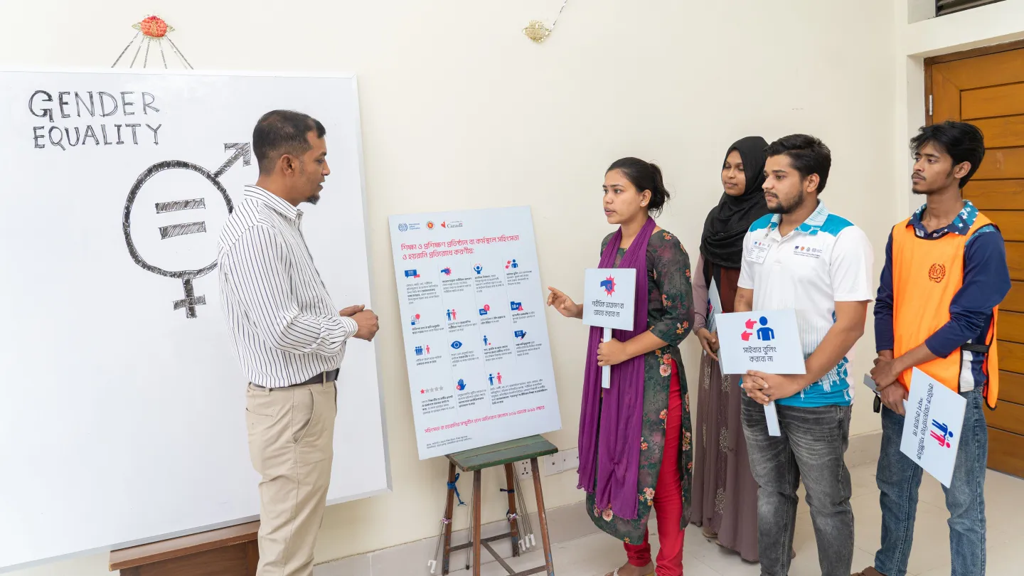 M. Ziaur Rahman se tient devant un tableau blanc sur lequel est inscrit « Égalité de genre ». Il s'adresse à un groupe de stagiaires qui tiennent des pancartes et des supports visuels illustrant différents aspects de l'égalité de genre et de l'inclusion.