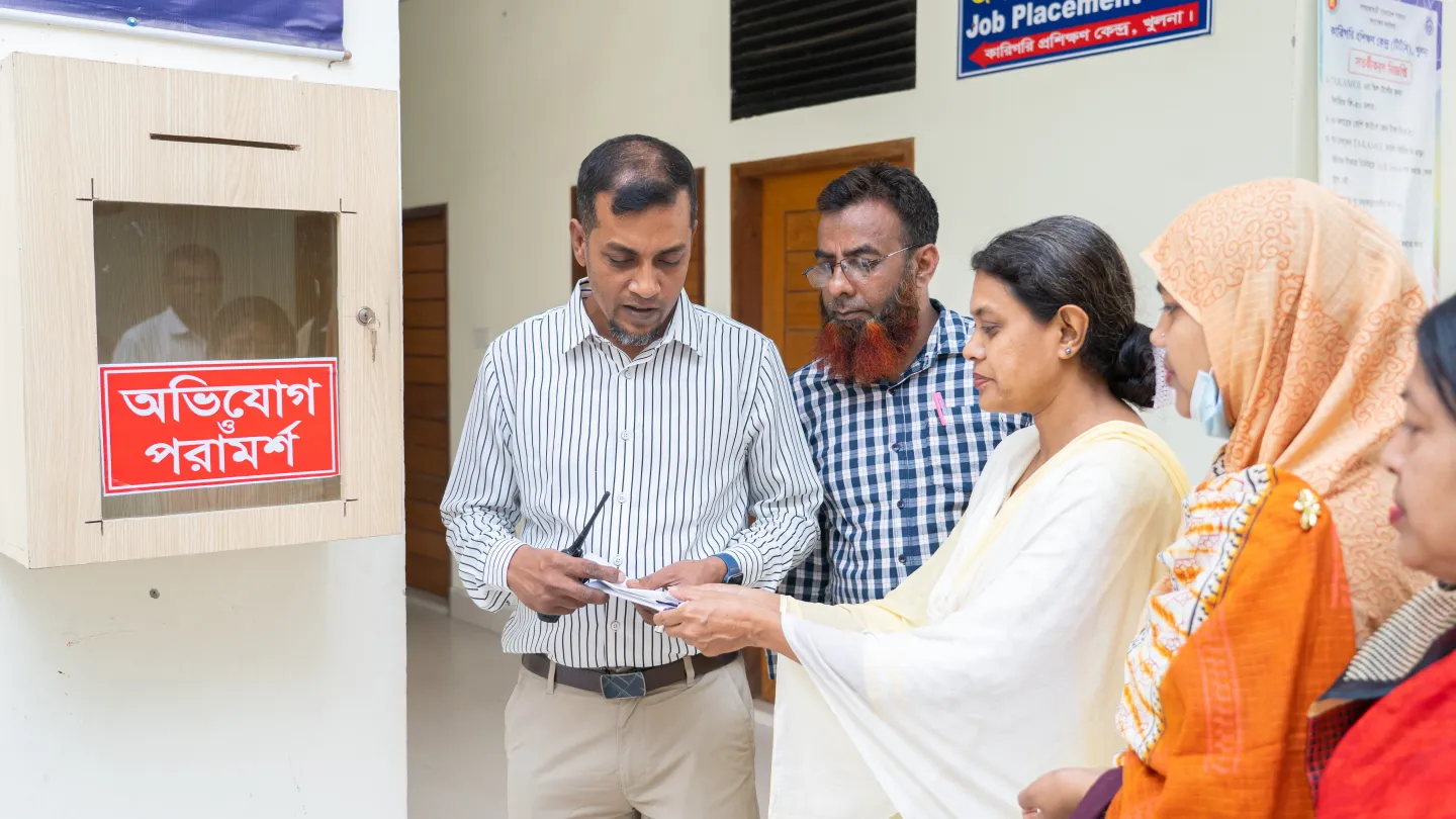 Md. Ziaur Rahman et des membres du Comité chargé des questions de genre du centre de formation – trois femmes et un homme – se tiennent à côté d’une boîte à suggestions et à réclamations. Ils examinent une note écrite sur une feuille de papier.