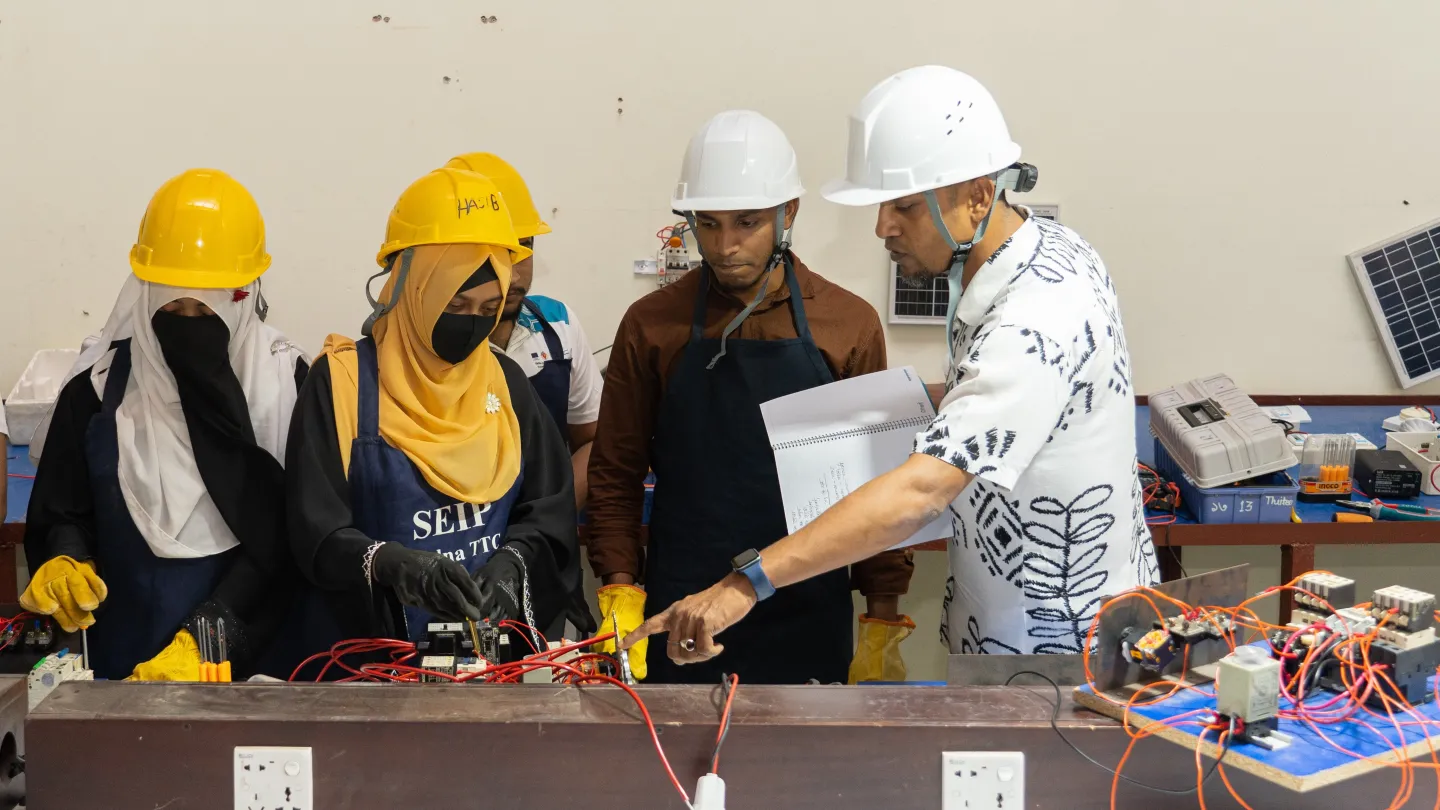 Md. Ziaur Rahman désigne un tableau d’installation électrique lors d’une séance de formation pratique. Deux stagiaires femmes et deux stagiaires hommes l’observent. Tous portent des casques de sécurité.
