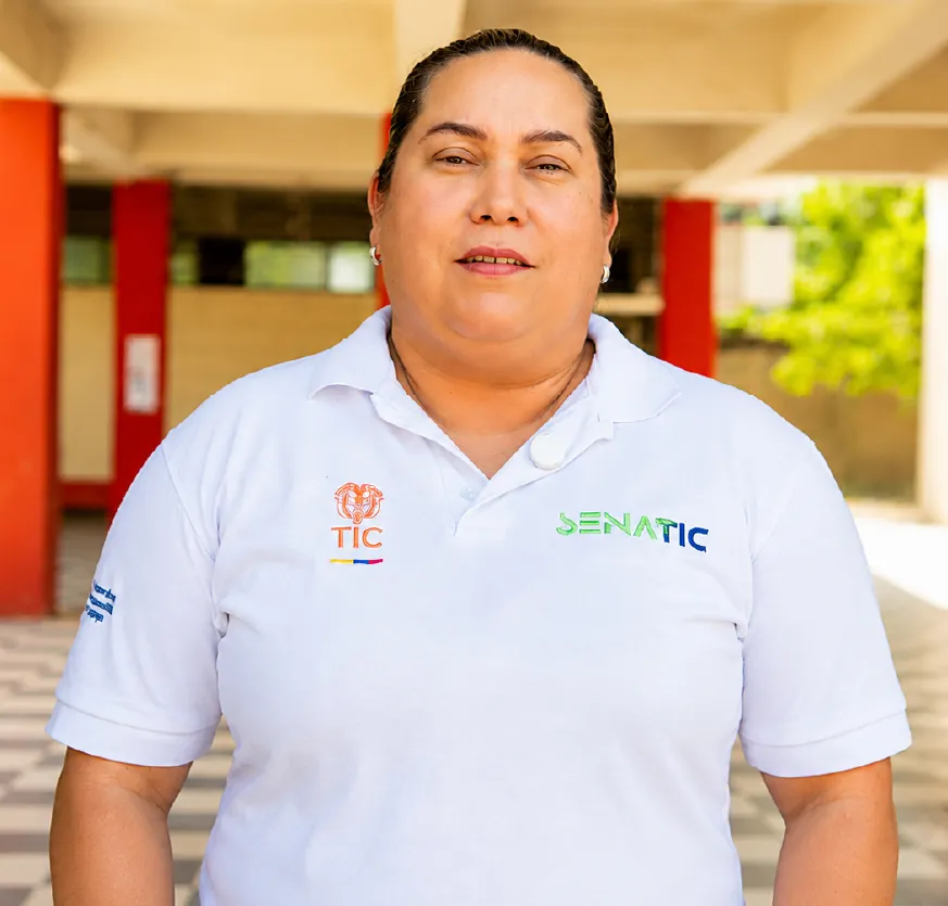 Una mujer se encuentra de pie en un corredor de una institución educativa. Viste una camiseta blanca del programa y mira hacia la cámara. Al fondo se observan columnas, vegetación y espacios abiertos del centro educativo.