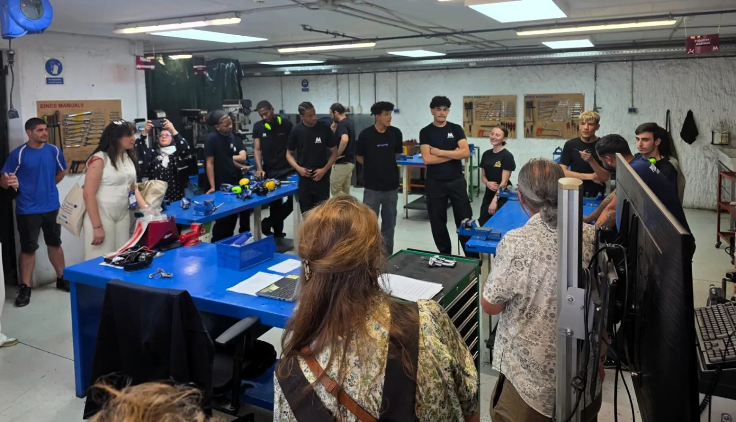 A group of visitors observes young trainees and instructors in a technical workshop equipped with workbenches and tools. Participants discuss training methods and skills development practices.