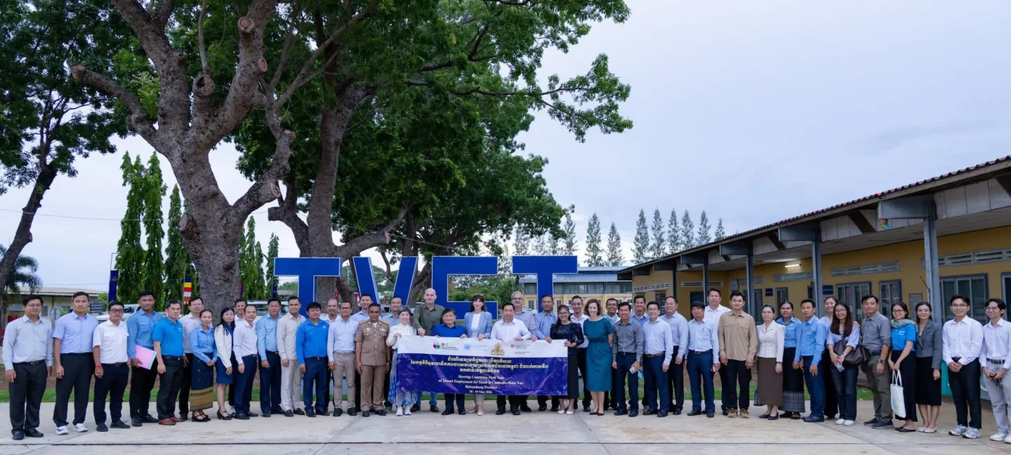A large group of people stand outdoors in front of a building, gathered around a banner for a group photo.