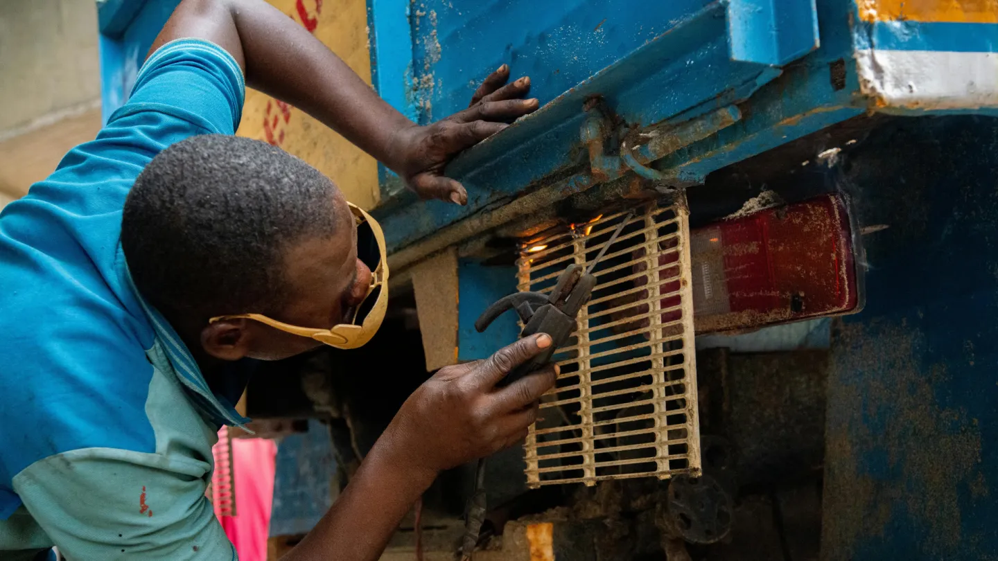 A mechanic repairs the rear electrical wiring of a blue tractor, using pliers to work near a tail light while leaning underneath the vehicle.