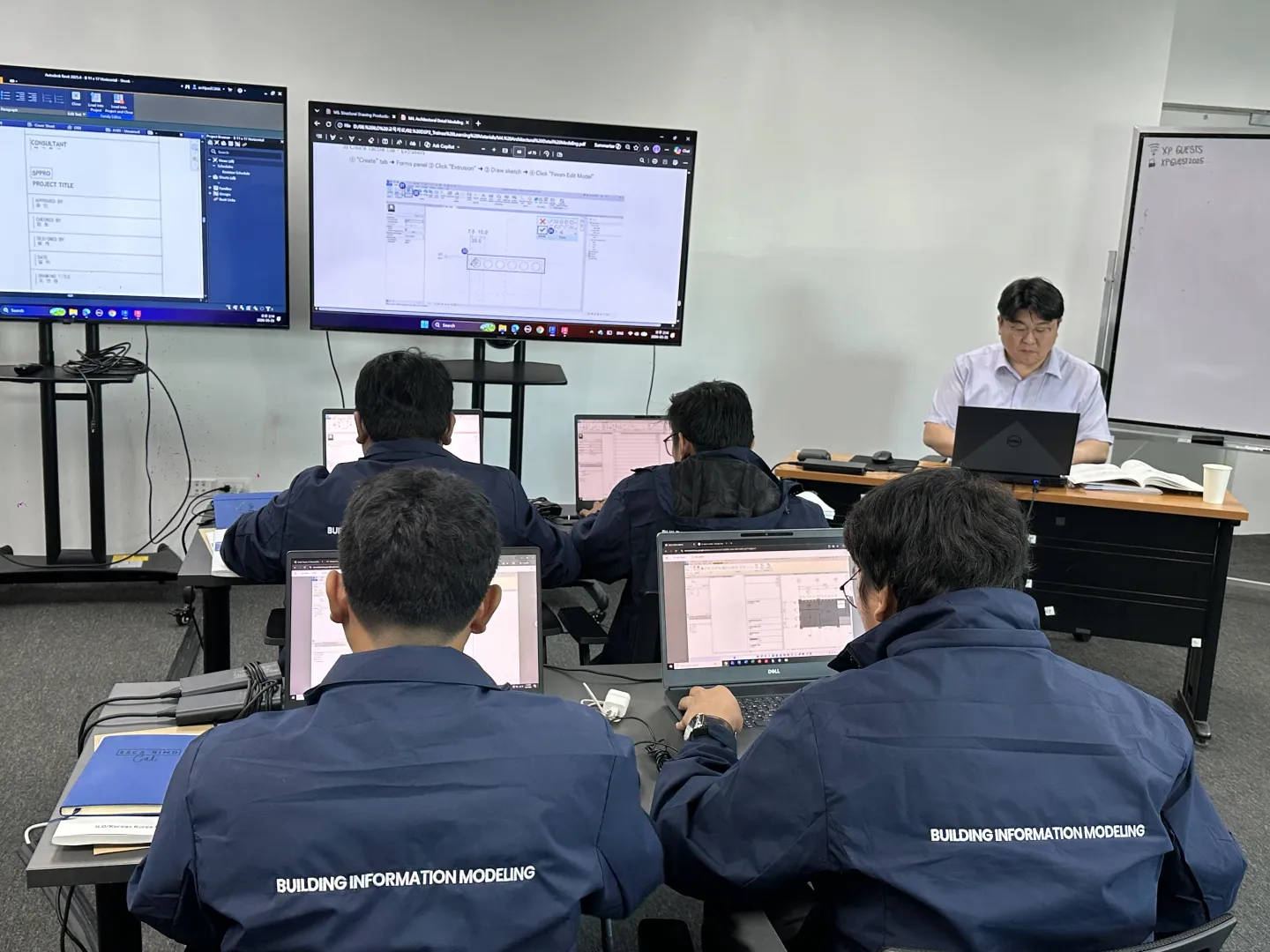 Participants use laptops during a Building Information Modeling (BIM) training session while an instructor demonstrates BIM software on large screens.