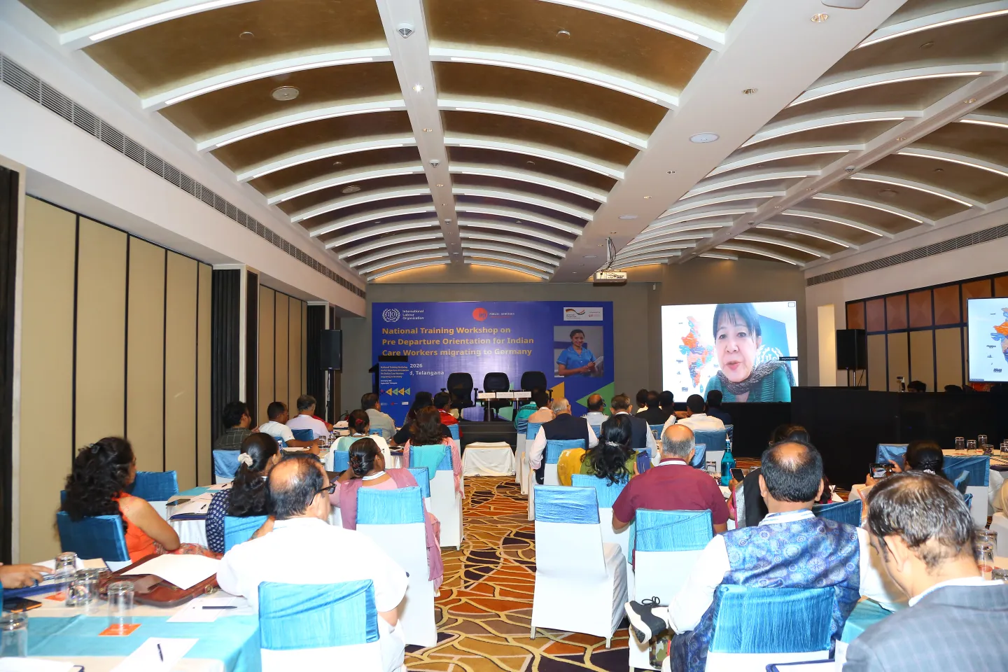 Participants facing a stage and large screen displaying a remote speaker via video conference.