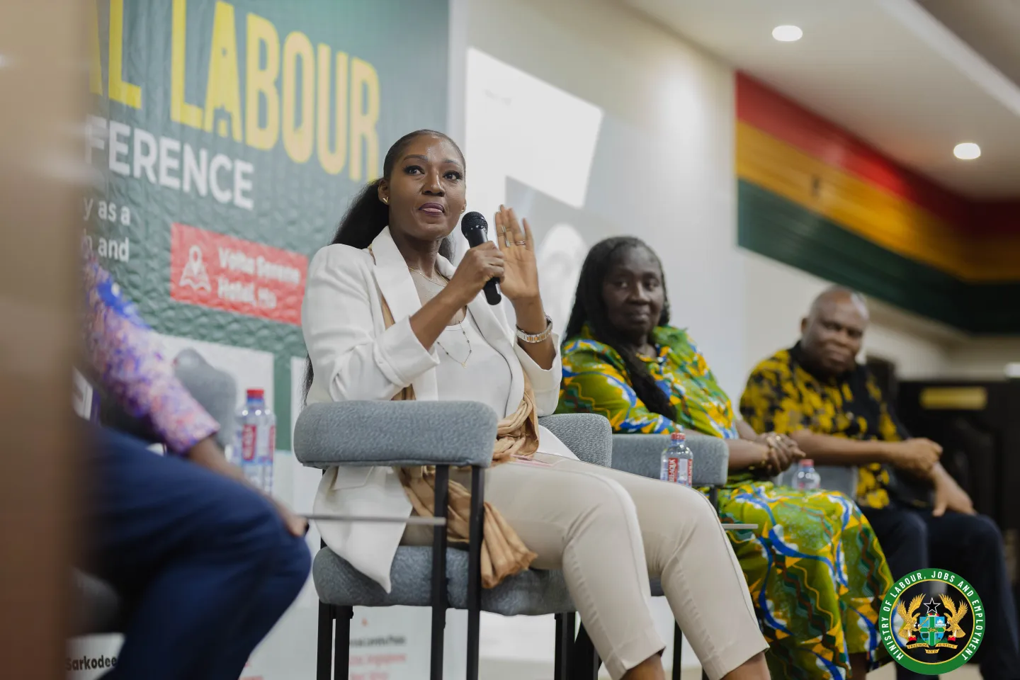 A woman seated on a panel speaks into a handheld microphone while other panelists listen.