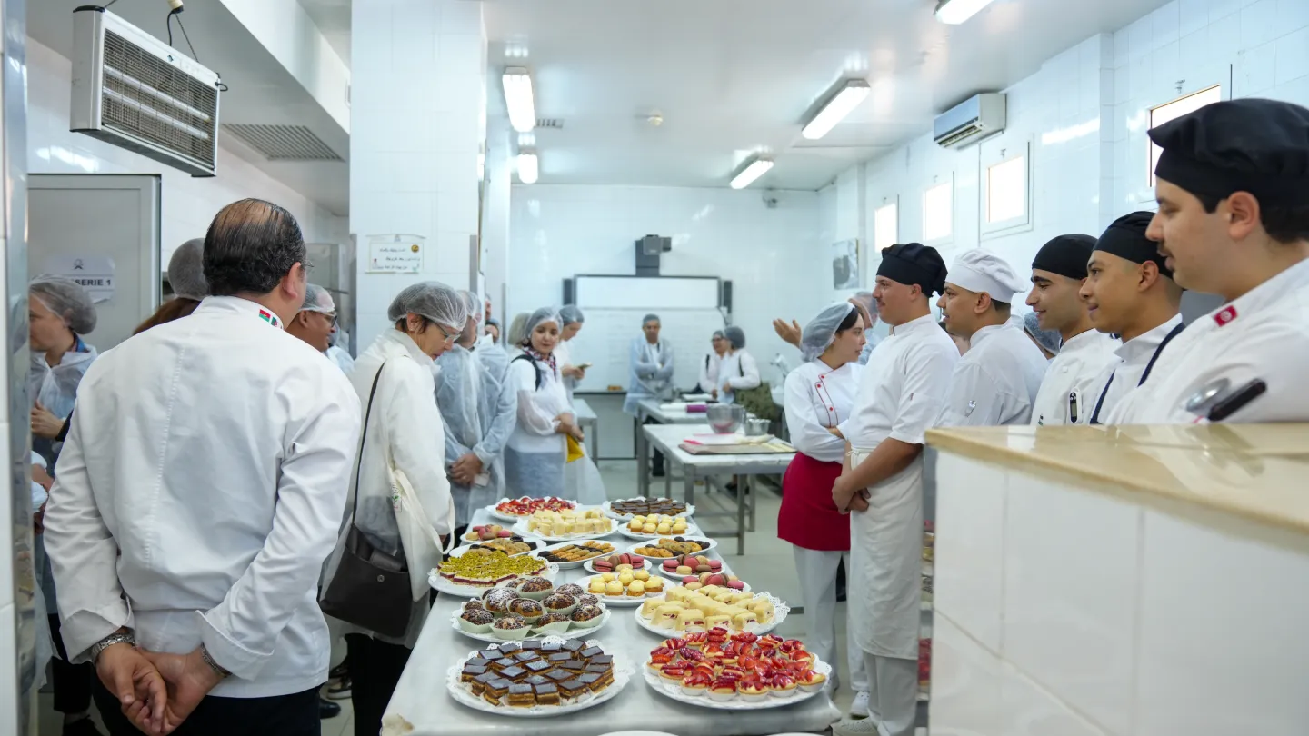 Des étudiants en tenue de cuisine présentent une sélection de pâtisseries et de desserts à des visiteurs dans une cuisine pédagogique. Plusieurs participants, équipés de charlottes et de blouses de protection, échangent avec les étudiants autour des préparations exposées.