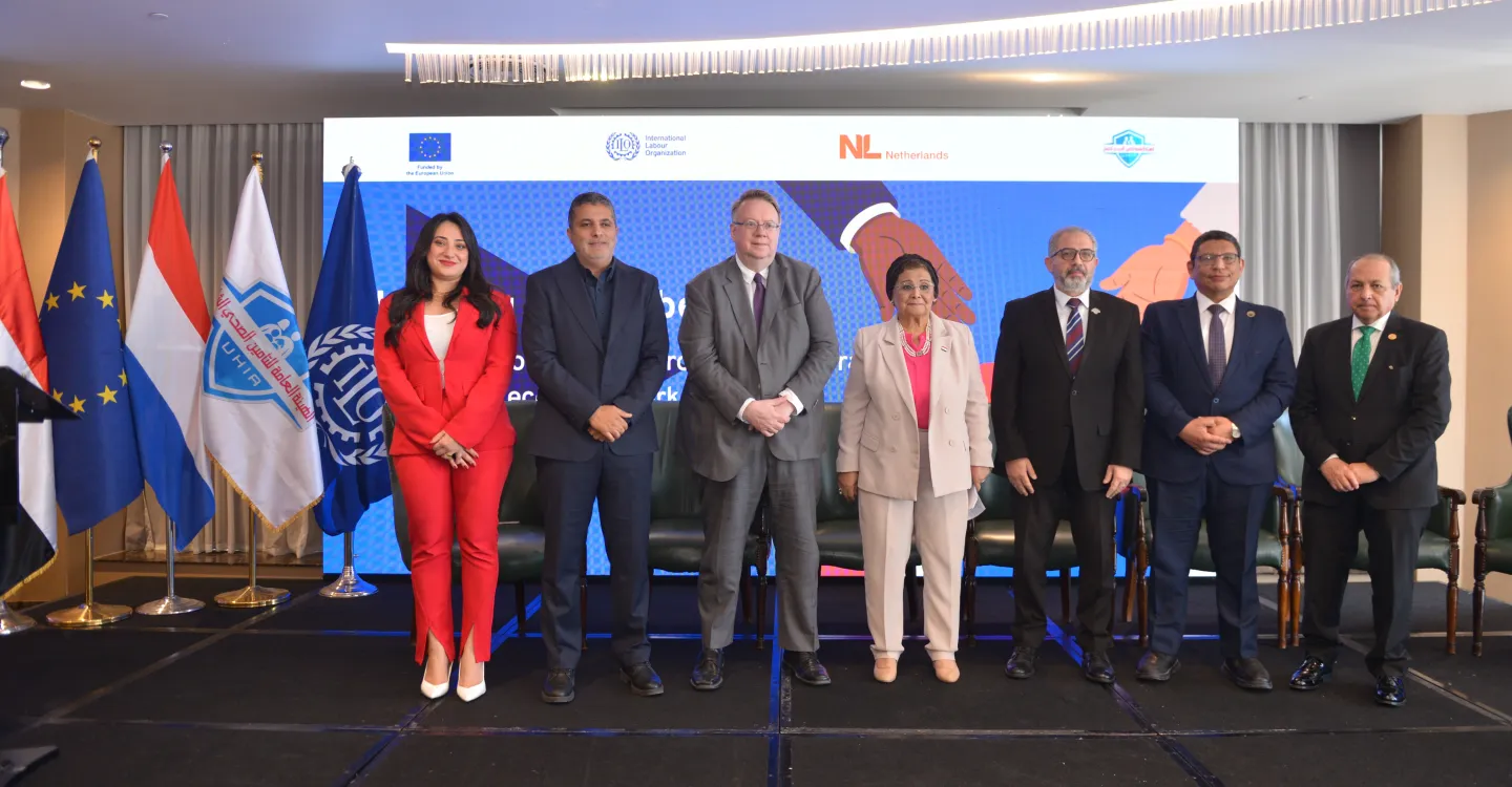 Seven individuals stand next to each other on a stage with the flags of Egypt, the EU, Netherlands, UHIA and the ILO next to them. Behind them is a blue backdrop on extending social protection for all.