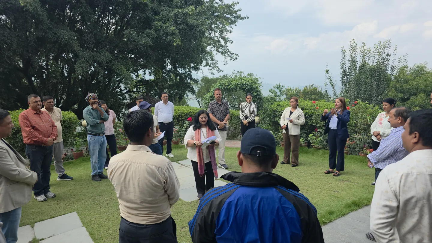 A facilitator addresses a group of participants standing in a circle on a garden lawn.